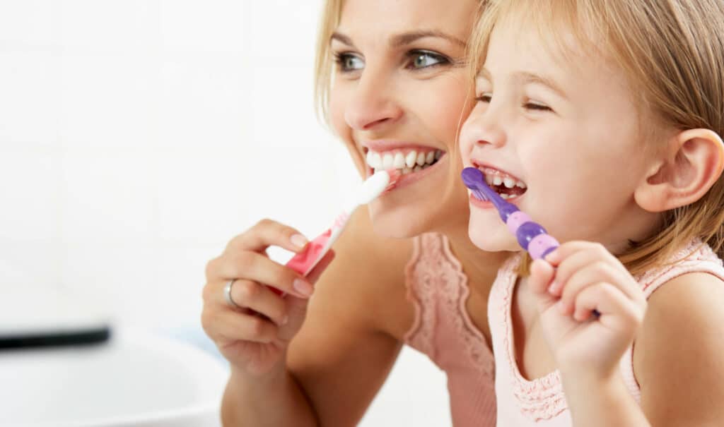 A person and child happily brush their teeth together in a bathroom, smiling brightly and focusing on their dental hygiene.