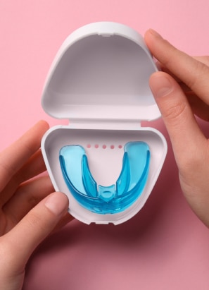 A person holds a white container with a blue dental mouth guard inside, against a pink background. The container is open, showing the guard clearly.
