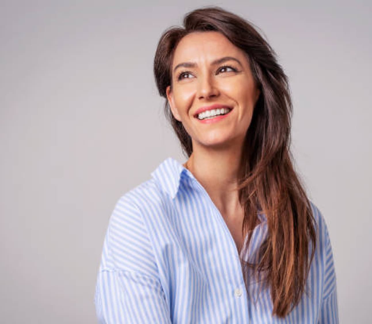 A person with long hair, wearing a blue and white striped shirt, smiles while looking to the side, against a plain gray background.