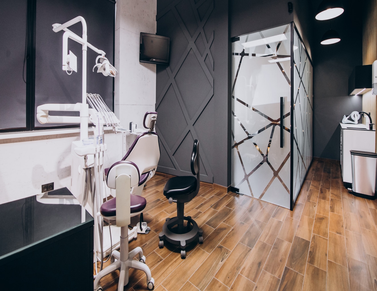 A modern dental clinic features a dental chair, equipment, wooden flooring, glass partitions, and sleek black and white decor. There are no people present.