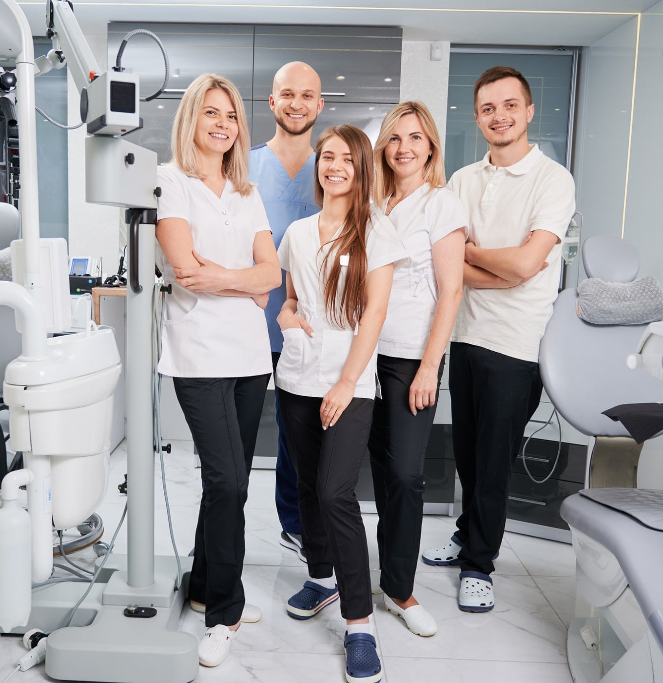 Five persons in dental attire stand in a well-equipped dental clinic, smiling and posing together for a group photo.