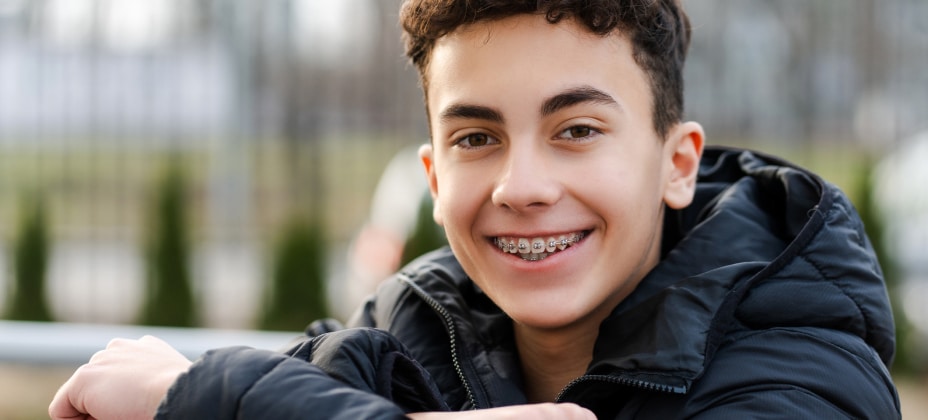 A smiling individual with braces is dressed in a black jacket, posing outdoors with blurred trees and a fence in the background.