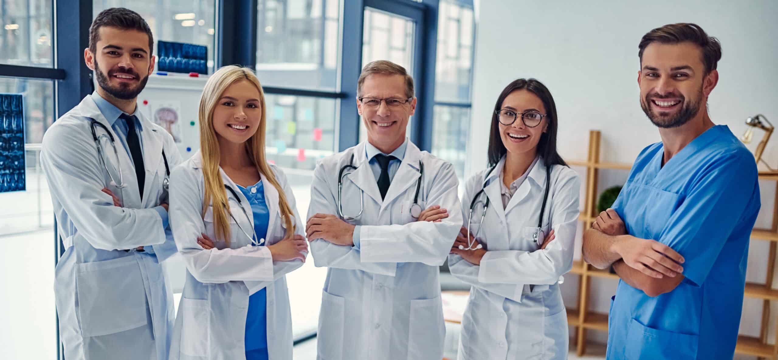 Five people in medical attire stand smiling with arms crossed in a modern, well-lit medical office or hospital setting, large windows in the background.